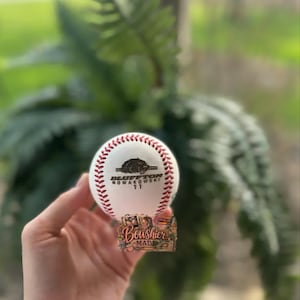 May include: A white baseball with red stitching. The ball features a logo with the text "Bluffton Nowakowski 11" and a bear graphic. The bottom of the ball has a decorative sticker with the text "Boushier Made".