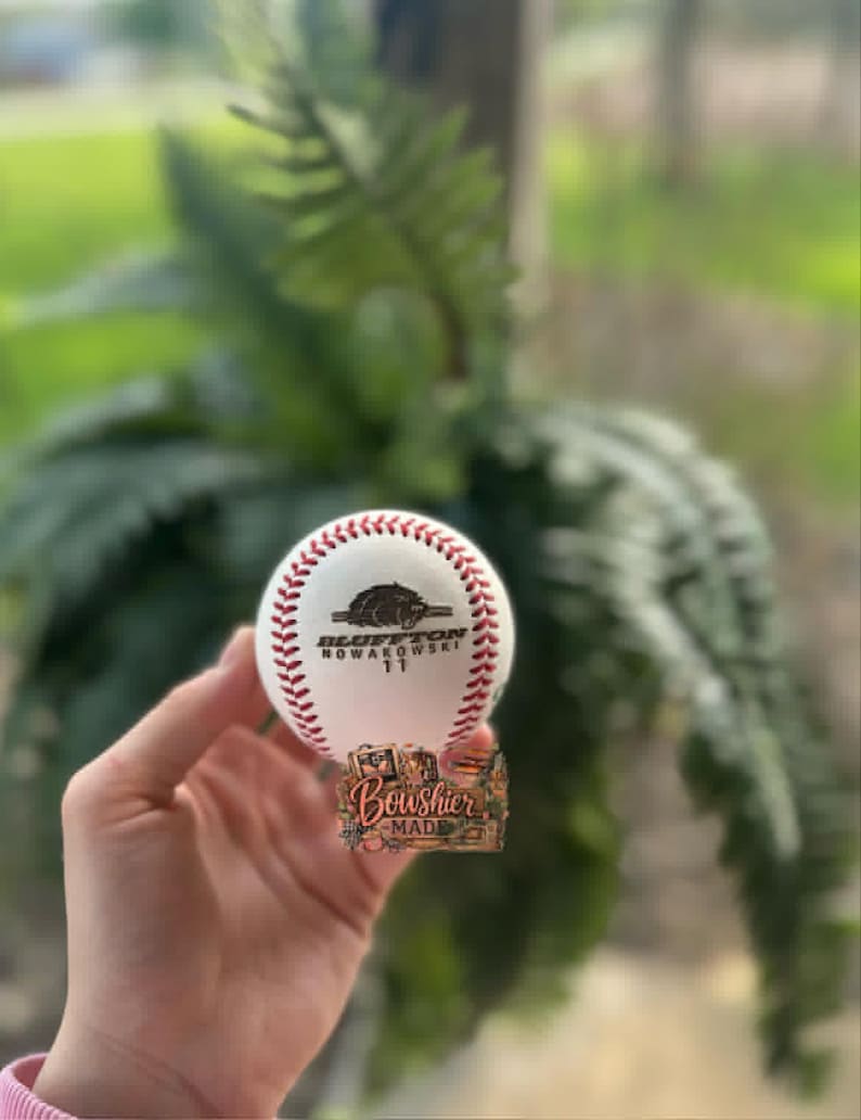 May include: A white baseball with red stitching. The ball features a logo with the text "Bluffton Nowakowski 11" and a bear graphic. The bottom of the ball has a decorative sticker with the text "Boushier Made".