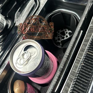 Puede incluir: Un portavasos rosa para coche con un borde morado que sostiene una lata plateada. Se ve el interior del coche, incluyendo un inserto para portavasos con estampado de vaca en blanco y negro y un letrero de madera que dice "Bowshier Made".