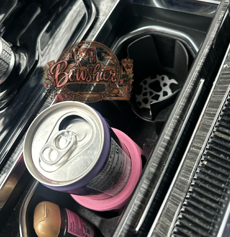 Puede incluir: Un portavasos rosa para coche con un borde morado que sostiene una lata plateada. Se ve el interior del coche, incluyendo un inserto para portavasos con estampado de vaca en blanco y negro y un letrero de madera que dice "Bowshier Made".