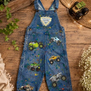 May include: Blue denim overalls featuring a Monster Jam logo on the chest pocket, multiple monster truck graphics, and colorful paint splatter accents. The pant legs have a black and white checkered pattern at the cuffs and adjustable straps.