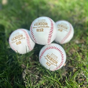May include: Four white baseballs with red stitching are displayed on green grass. Each ball is personalized with text, including "2026 SENIOR" and a name and number. The baseballs are likely commemorative items or gifts.
