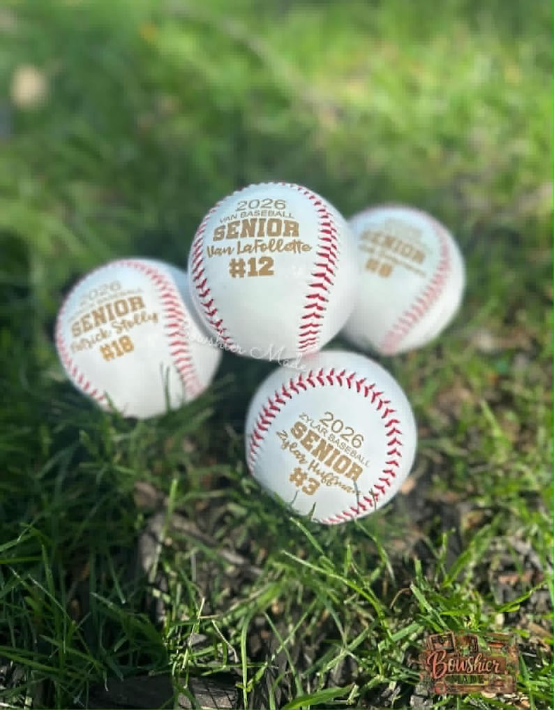 May include: Four white baseballs with red stitching are displayed on green grass. Each ball is personalized with text, including "2026 SENIOR" and a name and number. The baseballs are likely commemorative items or gifts.