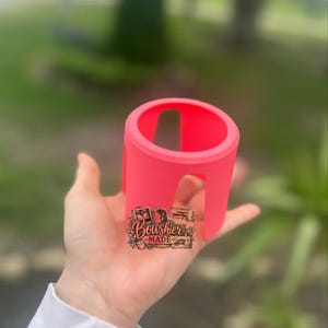 Puede incluir: Un portavasos rosa brillante, de forma cil&iacute;ndrica con un dise&ntilde;o recortado. El soporte se sostiene en una mano, con una peque&ntilde;a pegatina que dice "Boushier Made". El fondo est&aacute; borroso, con follaje verde.