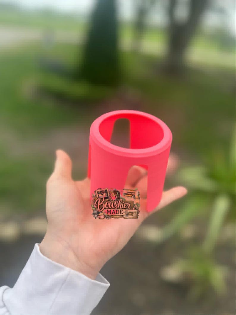 Puede incluir: Un portavasos rosa brillante, de forma cil&iacute;ndrica con un dise&ntilde;o recortado. El soporte se sostiene en una mano, con una peque&ntilde;a pegatina que dice "Boushier Made". El fondo est&aacute; borroso, con follaje verde.