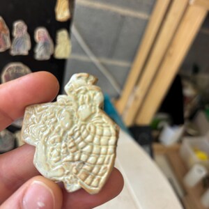 Peut inclure: Un pendentif en céramique crème en forme de croix avec un relief d'un couple. Le pendentif a une surface texturée et un motif quadrillé sur les vêtements de l'homme. D'autres pendentifs similaires sont visibles en arrière-plan.