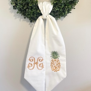 May include: A circular green boxwood wreath adorned with a white fabric ribbon tied in a knot at the bottom. The ribbon features an embroidered orange monogram letter H on the left side and an embroidered orange and green pineapple on the right.