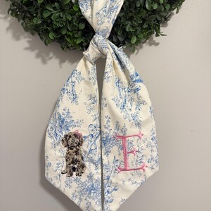 May include: A cream-colored fabric scarf featuring a blue toile de jouy pattern. The scarf is tied in a knot and displays an embroidered brown dog with a small pink bow on its head on one side and a large, decorative pink letter E on the other.