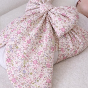 May include: A baby swaddle blanket with a large bow, featuring a delicate floral pattern in pink, white, and green. The bow is tied at the top, creating a soft, decorative element. The blanket is designed for newborns.