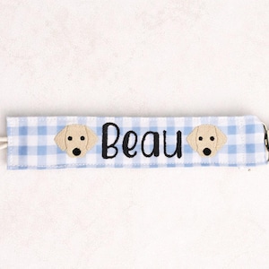 May include: A light blue and white gingham fabric pacifier clip featuring the embroidered name Beau in black script, flanked by two tan dog face appliques, finished with a white elastic loop on one end and a silver metal clip on the other.
