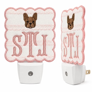Könnte beinhalten: Ein weißes Nachtlicht mit einer quadratischen Stoffabdeckung mit gewellten rosa Rändern. Der Stoff hat ein hellrosa Streifenmuster, ein gesticktes braunes Französische Bulldogge-Gesicht und die gestickten rosa Buchstaben STI.