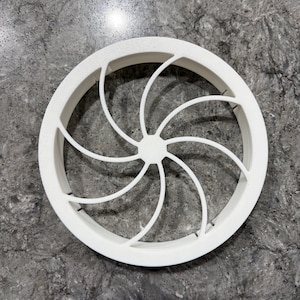 May include: A circular white plastic component featuring a central hub with eight curved, radiating spokes that connect to an outer ring frame. The design creates a symmetrical, fan-like pattern with open spaces between each of the curved structural supports.