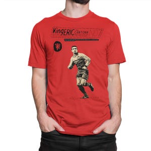 Puede incluir: Camiseta roja con un gráfico en blanco y negro de un futbolista en acción. La camiseta incluye el texto "King Eric Cantona No. 7" y un logotipo del equipo. El diseño es de estilo vintage.