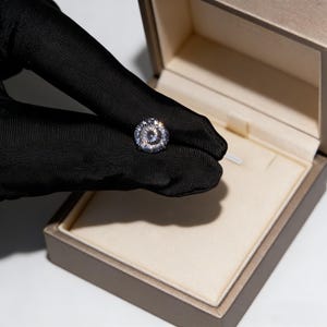 May include: A sparkling, round-cut diamond held by a gloved hand, presented in an open jewelry box. The diamond's facets reflect light, creating a brilliant display. The box is a neutral color, with a soft, cream-colored interior.