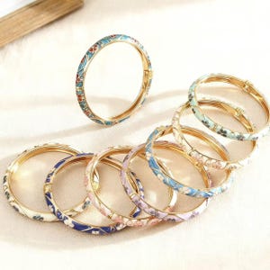 May include: A selection of gold-coloured, hinged bangles with vibrant floral enamel designs. The bracelets showcase diverse colour schemes, including blue, red, white, and purple, with detailed patterns. The bangles are presented on a light-coloured surface.