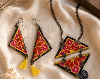 Op de afbeelding: Een sieradenset bestaande uit twee driehoekige oorbellen en een bijpassende ketting. Elk item is gemaakt van zwarte stof met levendige roze en gele geometrische borduursels, antiek bronzen metalen accenten en kleine gele draadkwastjes.