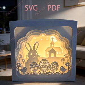 Peut inclure: Boîte à ombre sur le thème de Pâques en papier découpé, avec un lapin, des œufs de Pâques, une église et des fleurs. La boîte est éclairée de l'intérieur, créant une lueur chaleureuse. Le texte "SVG PDF" est visible.
