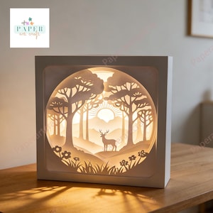 May include: A white, square paper art light box depicting a forest scene. The illuminated interior shows a deer, trees, and a sun. The box has the text "PAPER art crafts". The light box is on a wooden surface.