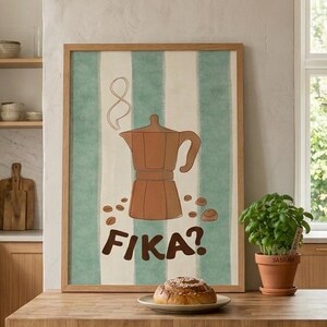 May include: A framed poster with a brown coffee maker illustration and the word "FIKA?" against a green and white striped background. A plate with a pastry sits on a wooden countertop. A potted plant is next to the plate.