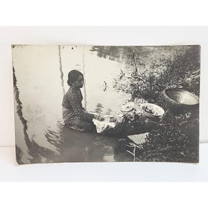 May include: Vintage black and white photograph depicting a person washing clothes in a body of water. The person is seated, wearing patterned clothing, and surrounded by laundry and a woven basket. The scene is set outdoors.
