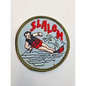 May include: Embroidered patch featuring a water skier in red shorts and a black top, with the word "SLALOM" in red letters. The patch has a light blue background and a green border, depicting water and a skier.