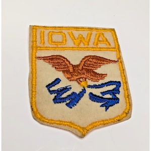 Puede incluir: Un parche bordado en forma de escudo con fondo color crema y borde amarillo. La parte superior presenta la palabra IOWA en hilo amarillo. Debajo, un águila marrón aparece sobre un diseño de cinta azul, todo bordado sobre la tela.