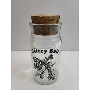 Puede incluir: Un tarro de cristal transparente con tapón de corcho, etiquetado "Celery Salt" en escritura negra. Una ilustración en blanco y negro de tallos y raíces de apio está debajo del texto. El tarro está lleno de un polvo blanco.