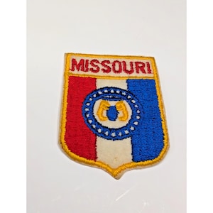 May include: A shield-shaped embroidered patch featuring the word MISSOURI in red text at the top. The design includes vertical red, white, and blue stripes with a central circular emblem containing a blue ring and yellow details on a white background.