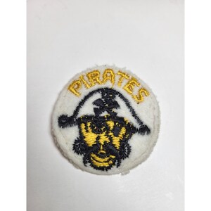 May include: Round, white embroidered patch with the word "PIRATES" in gold thread. Below the text is a black and gold pirate-themed design. The patch is likely for a jacket or bag.