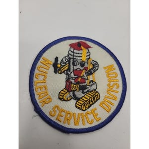 May include: A circular patch with a cartoon robot design. The robot is gray, yellow, and red, wearing a graduation cap. The patch has the words "NUCLEAR SERVICE DIVISION" in yellow text around the edge, with a blue border.