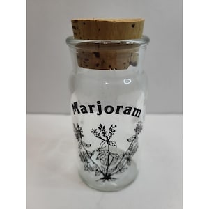 Puede incluir: Un tarro de vidrio transparente con tapón de corcho, etiquetado "Marjoram" en escritura negra. El tarro presenta una ilustración botánica en blanco y negro de plantas de mejorana. El tarro está diseñado para almacenar hierbas y especias.