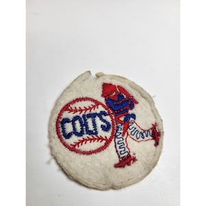 May include: Vintage Colts baseball patch. The patch is round and features a baseball with the word "COLTS" in blue lettering. A baseball player in red and blue uniform is running. The patch is made of a cream-colored fabric.