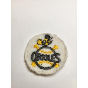 May include: A round, white patch featuring the word "Orioles" in black lettering, with a baseball design. The patch includes a yellow and black bee wearing a hat. The baseball design has yellow stitching and crossed yellow bats.
