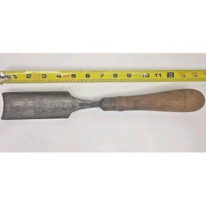 May include: A vintage woodworking chisel with a wide, flat blade and a wooden handle. The metal blade shows signs of wear and age. The handle is a light brown color. The chisel is approximately 12 inches long.
