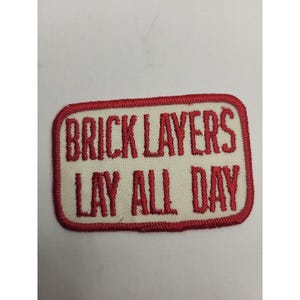 Könnte beinhalten: Rechteckiger Aufnäher mit weißem Hintergrund und rot gesticktem Text und Rand. Der Text lautet "BRICK LAYERS LAY ALL DAY". Der Aufnäher ist wahrscheinlich für Arbeitskleidung oder eine Uniform.