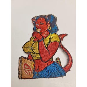 Puede incluir: Parche bordado que representa a una mujer diablo de piel roja con pelo azul, cuernos y cola. Lleva una camisa amarilla y vaqueros azules, sosteniendo una caja con un logotipo. El parche está detallado con varios colores y texturas.
