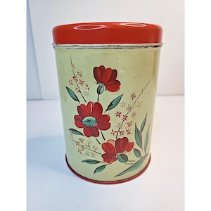 May include: Vintage metal canister with a red lid and base. The canister has a light green background and is decorated with red flowers, green leaves, and white accents. A decorative kitchen storage container.