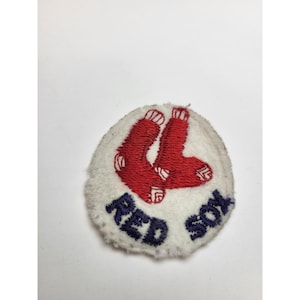 May include: A circular patch with the words "RED SOX" in blue. Two red socks are embroidered on a white, fuzzy background. The patch is a sports-themed emblem.