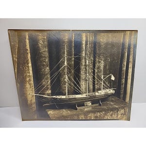 May include: Sepia-toned photograph of a detailed model ship, a tall sailing vessel with multiple masts and rigging. The model is displayed on a dark surface, with a dark, textured backdrop. A small white flag is visible on the ship.