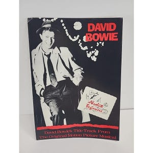 Può includere: Copertina di un album vintage con una foto in bianco e nero di David Bowie. Lo sfondo è nero con il nome "DAVID BOWIE" in rosso. Il titolo dell'album "Absolute Beginners" è anch'esso visibile. Il testo in basso recita "David Bowie's Title Track From The Original Motion Picture Musical."