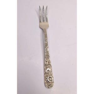 May include: A silver-toned metal fork featuring three tines and an ornate handle decorated with a detailed embossed floral pattern. The handle displays various flower shapes and leaves in a relief design that extends from the base to the neck.