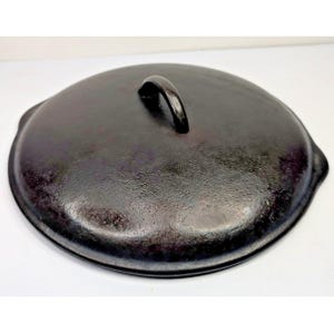 May include: A round, black cast iron skillet lid with a textured surface and a central arched handle. The lid features a slightly raised rim and two small, integrated side tabs for lifting, showcasing a dark, seasoned metallic finish throughout.