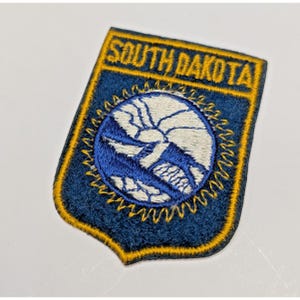 May include: A shield-shaped embroidered patch with a dark blue background and a yellow border. The text SOUTH DAKOTA is stitched in yellow at the top. A central circular emblem features a white sun and blue landscape design with yellow rays.