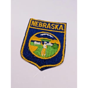 May include: A shield-shaped embroidered patch featuring a deep blue background with a yellow border. The word NEBRASKA is stitched in yellow at the top. The center displays a circular scene with a person in a field, hills, and a sky.