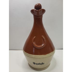 May include: A brown ceramic decanter with a rounded body and a narrow neck, topped with a small, round stopper. The decanter features a curved handle and the word "Scotch:" in black lettering on the lower portion.