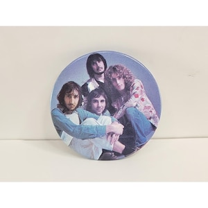 Puede incluir: Un botón redondo con una foto de una banda de rock. La imagen muestra a cuatro hombres con pelo largo y ropa colorida. El fondo es de color azul claro.