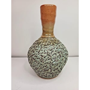 Puede incluir: Un jarrón de cerámica artesanal con un cuerpo texturizado y estampado en tonos verdes y marrones. El jarrón tiene un cuello estrecho y una base más ancha y redondeada. La parte superior es de un marrón cálido y terroso.