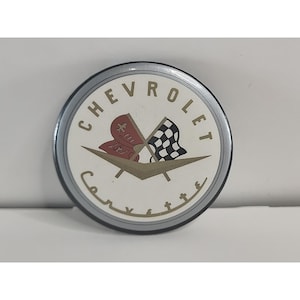May include: A round, silver and white Chevrolet Corvette emblem. The emblem features the word "Chevrolet" in a circular pattern, with "Corvette" in a cursive font. It includes a red flag and a checkered flag.