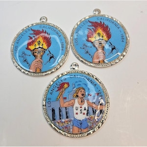 Op de afbeelding: Drie ronde, zilverkleurige hangers met het logo van de Olympische Spelen van Los Angeles 1984. Elke hanger heeft een lichtblauwe achtergrond met een fakkel en de tekst "LOS ANGELES CALIFORNIA - 1984". Eén hanger toont een hardloper.