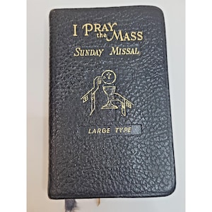 May include: A black, textured-cover Sunday missal book titled I PRAY the MASS. The cover features gold-colored lettering and a central gold graphic depicting a chalice and host. The text LARGE TYPE is printed in gold near the bottom of the cover.